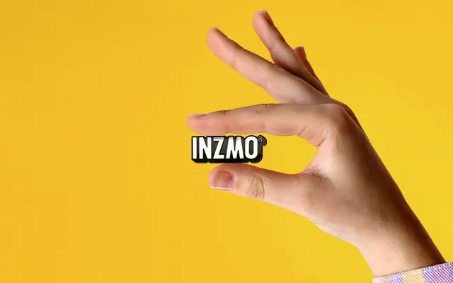 Inzmo enamel pin held between two fingers on yellow, photographic, minimal, vibrant