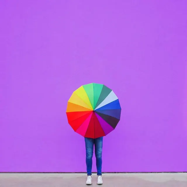 Person Holding a Rainbow Umbrella Against Purple Wall, minimal, playful, vibrant