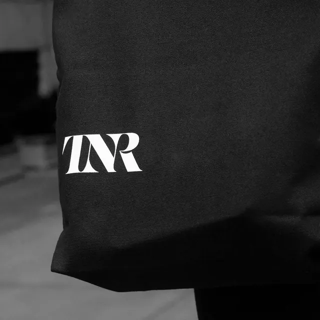 Close-up of dark fabric garment with white 'IVR' serif wordmark logo, minimalist, technical, monochrome