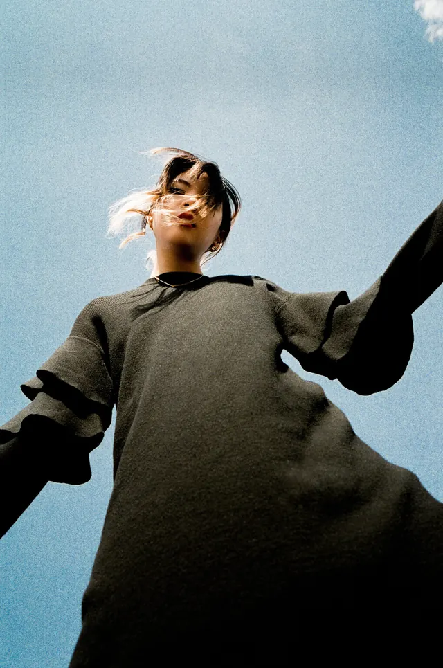 Person photographed from below against blue sky with outstretched arms, photographic, minimalist, cool