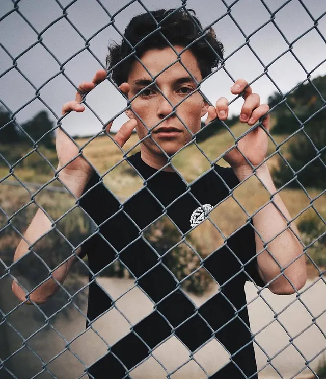 Portrait behind chain-link fence with hands gripping, photographic, minimal, cool