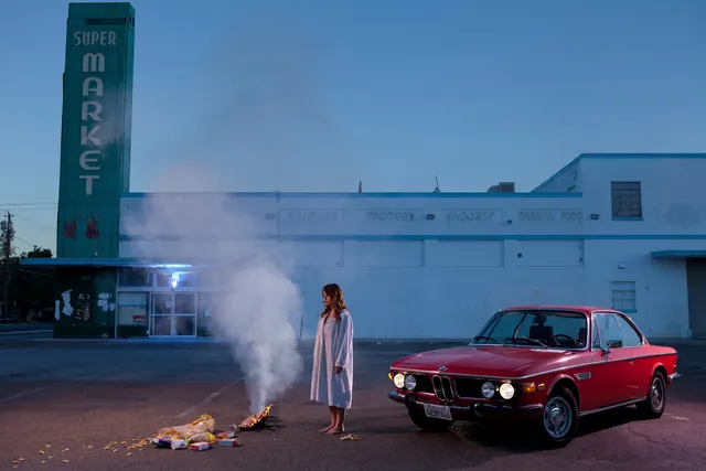 Woman in robe by burning groceries and vintage car, photographic, editorial, dark