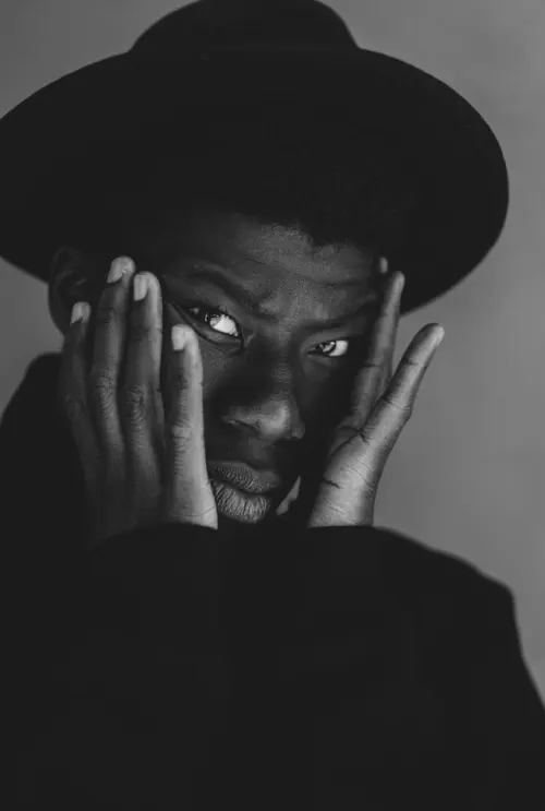 Intense portrait with wide hat and hands framing face, photographic, minimalist, dark