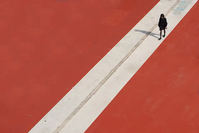 Solitary figure walking on white line across red plaza, minimal, photographic, vibrant