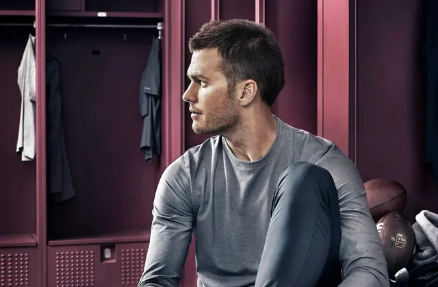 Tom Brady seated in maroon locker room, photographic, dark