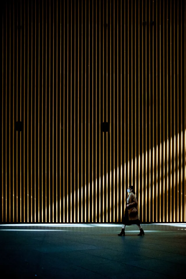 Woman walking past illuminated vertical slat wall at night, photographic, minimal, dark