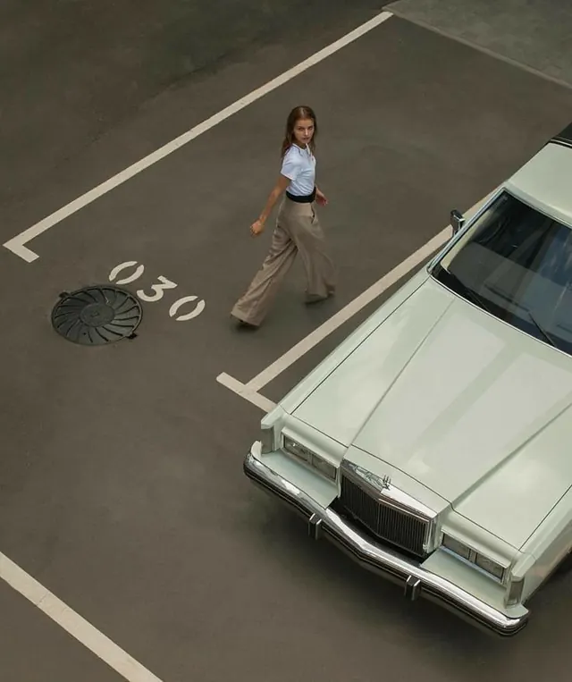 Overhead photograph of woman walking past parked vintage car with address marking, photographic, minimal, muted