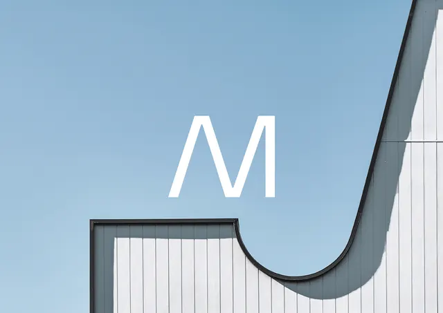 Large white M on blue sky with building facade, minimal, photographic, light