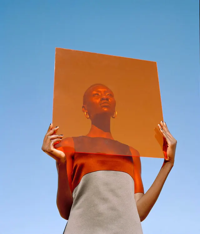 Model holding translucent orange panel against blue sky, editorial, photographic, vibrant