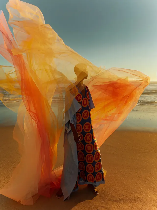 Person on beach with flowing orange sheer fabric, photographic, editorial, warm
