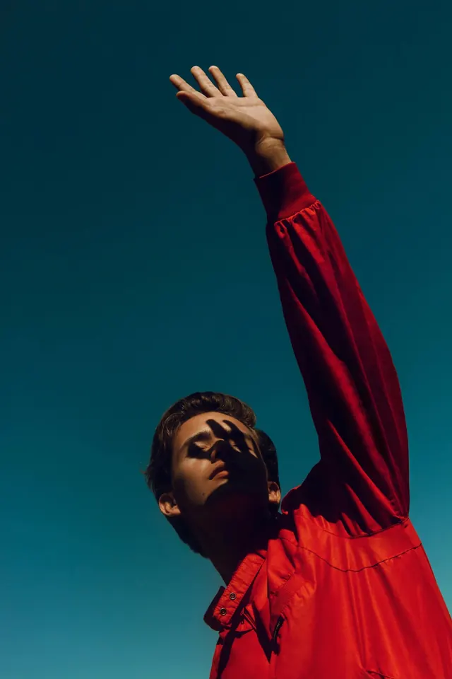Portrait with raised arm and bold red jacket against teal background, photographic, editorial, vibrant
