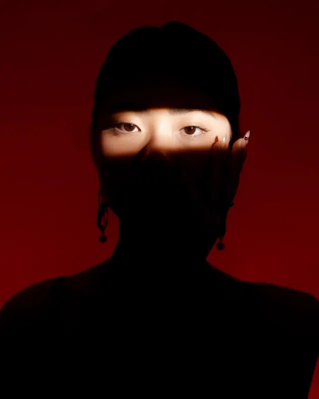 Silhouetted figure in black hood against deep red background with exposed eyes, photographic, minimal, dark