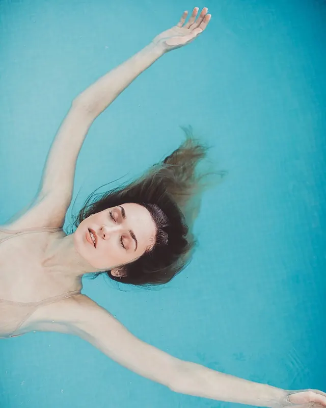 Woman floating in turquoise pool water, overhead view, photographic, minimal, vibrant