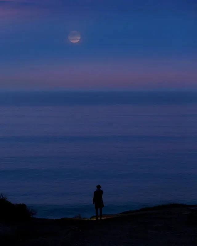 Silhouetted figure gazing at moonrise over ocean, photographic, dark-mode, dark